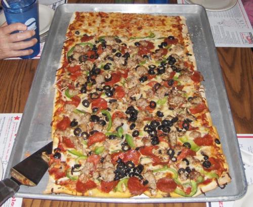 A large rectangular pizza topped with pepperoni, sausage, green peppers, and black olives on a metal tray.
