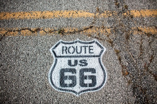 A painted Route 66 sign on cracked asphalt, featuring a black shield with white text.