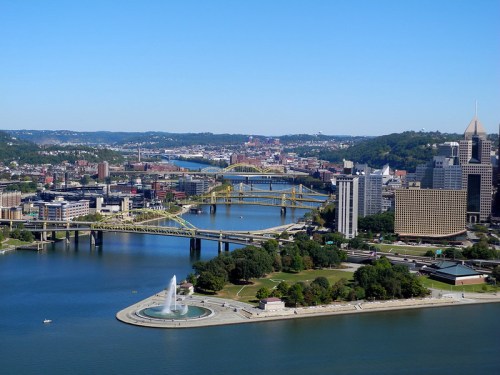 A panoramic view of Pittsburgh, featuring rivers, bridges, and city buildings under a clear blue sky.