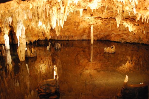 A stunning cave interior with stalactites and stalagmites reflected in still water, illuminated by warm lighting.