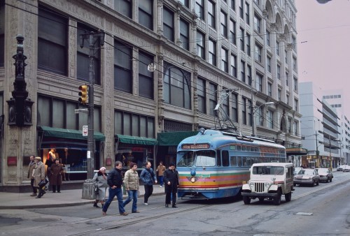 A colorful streetcar on a city street, with pedestrians and vintage cars near historic buildings.