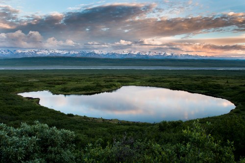A serene landscape featuring a calm pond reflecting clouds and mountains under a colorful sky. Lush greenery surrounds the water.