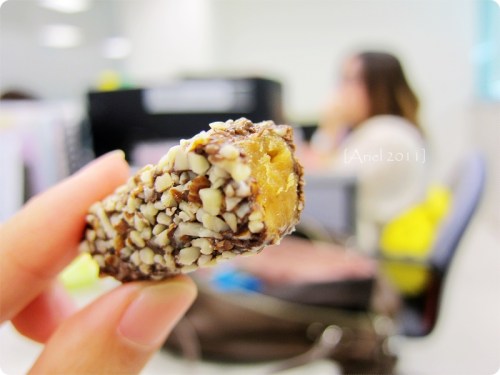 A close-up of a chocolate-covered snack with nuts, held in hand, with a blurred office background.