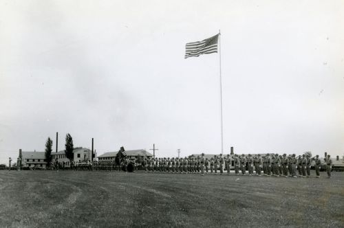 A black and white photo of soldiers in formation, with a large American flag flying in the background.