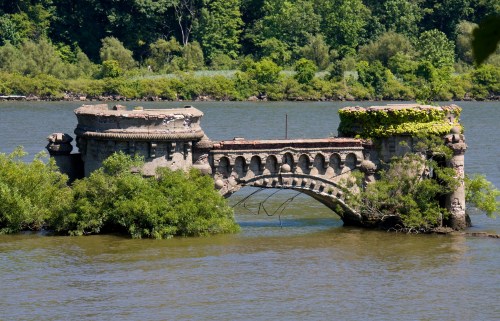Ruined stone bridge partially submerged in a river, surrounded by lush greenery and trees.