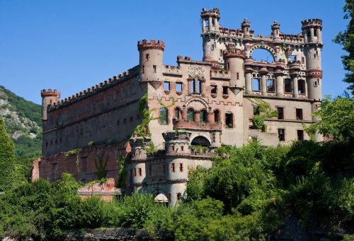 A large, crumbling castle with towers and arches, surrounded by greenery and set against a clear blue sky.