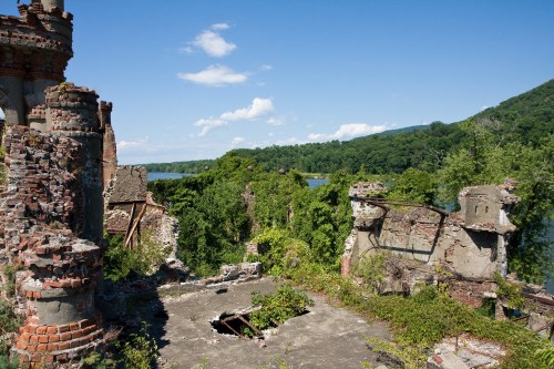 Ruins of a brick structure overgrown with vegetation, overlooking a river and green hills under a clear blue sky.
