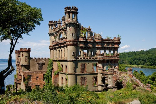 A crumbling, ornate castle ruin covered in greenery, overlooking a river with trees in the background under a clear blue sky.