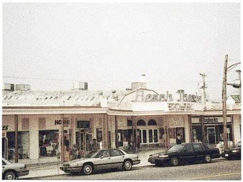 A vintage photo of a beachside shopping area with storefronts and parked cars in front.