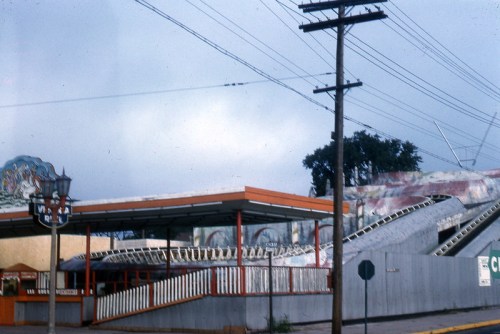 A vintage amusement park with a roller coaster, colorful murals, and power lines against a cloudy sky.