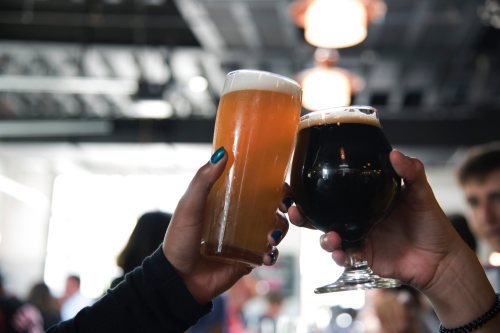 Two hands clink glasses of beer—one light and frothy, the other dark and rich—against a blurred background.