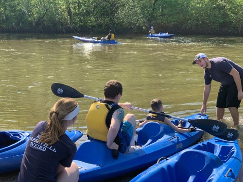 A group of people kayaking on a river, with one person assisting another in a blue kayak. Trees line the riverbank.