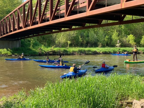 A group of kayakers paddles under a bridge on a calm river, surrounded by lush greenery.