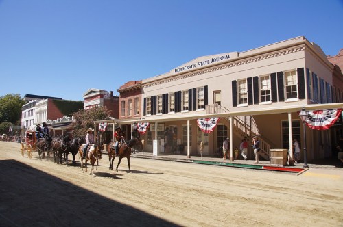 A historic street scene with horses and riders, flanked by buildings decorated with bunting and a clear blue sky.