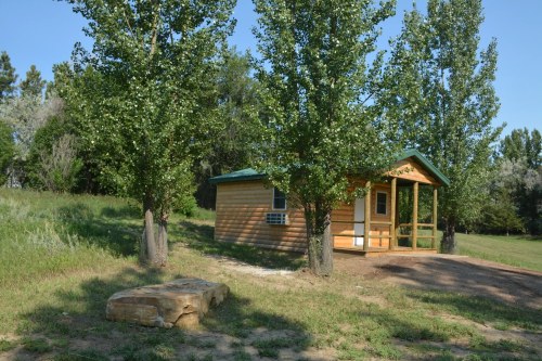 A small wooden cabin surrounded by trees, with a porch and a grassy area in front.