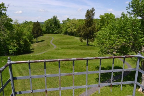 View from a tower overlooking a lush green landscape with winding paths and trees under a clear blue sky.