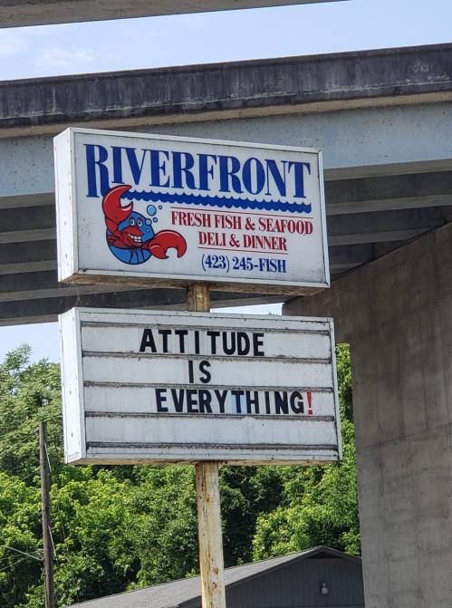 Sign for Riverfront Fresh Fish & Seafood reads "ATTITUDE IS EVERYTHING!" against a blue sky and greenery.