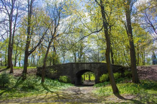 A stone bridge arches over a path surrounded by lush green trees and bright spring foliage.