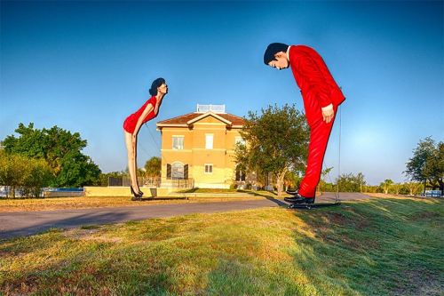 Giant sculptures of a man in a red suit and a woman in a red dress loom over a house in a grassy landscape.