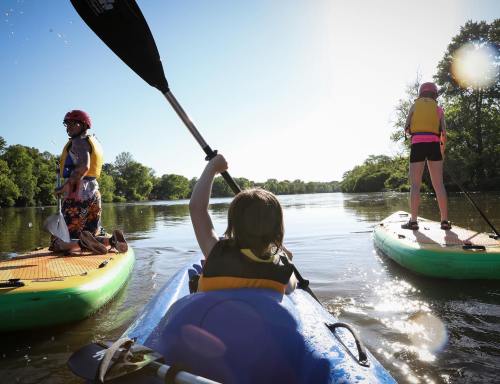 A child paddles a kayak while two adults on paddleboards enjoy a sunny day on a calm river.