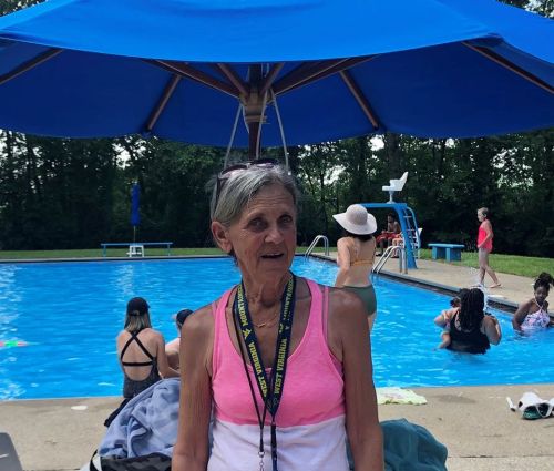 An older woman stands by a pool, wearing a lanyard, with people swimming and relaxing in the background under a blue umbrella.