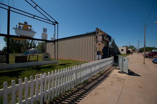 A sunny street scene featuring a metal building with colorful signs and a white picket fence in the foreground.