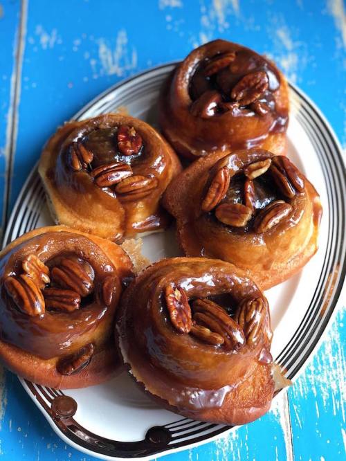 A plate of five sticky pecan cinnamon rolls with a glossy caramel glaze on a blue wooden surface.