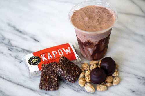A cup of chocolate smoothie next to a KAPOW protein bar, chocolate-covered snacks, and scattered nuts on a marble surface.
