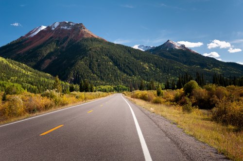 A scenic road stretches through a lush valley, flanked by mountains with patches of snow and vibrant greenery.