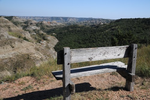 A weathered wooden bench overlooks a vast, rugged landscape of hills and valleys under a clear blue sky.