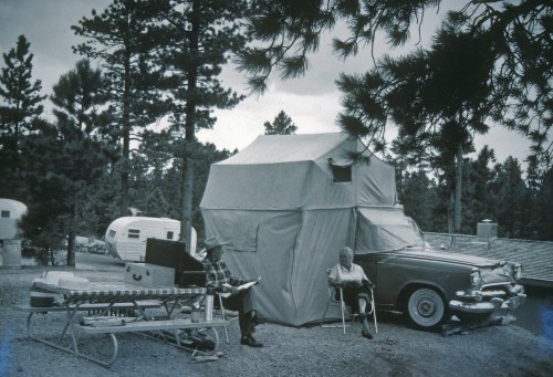A vintage camping scene with a car and tent, featuring two people relaxing outdoors among trees.