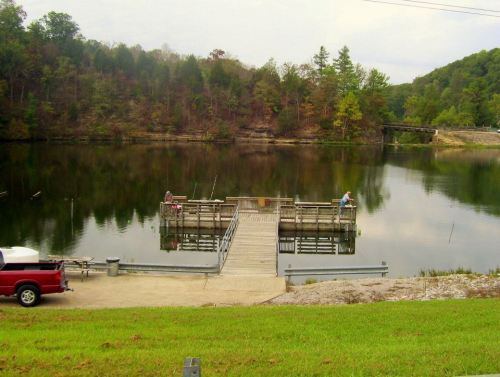A serene lake scene with a fishing dock, surrounded by trees and a grassy area, featuring two people fishing.