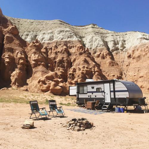 A camper parked in a desert landscape with colorful rock formations, two chairs, and a fire pit nearby.