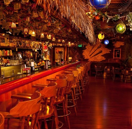 A vibrant bar interior with wooden stools, tropical decor, and colorful hanging lights, creating a lively atmosphere.