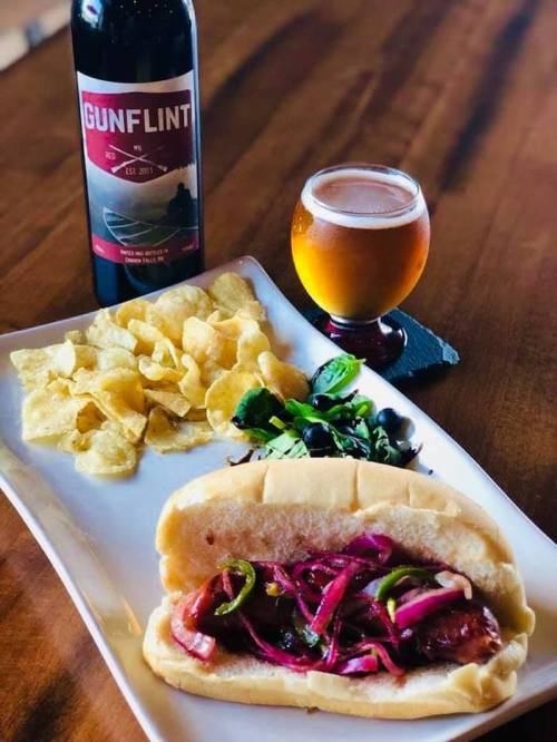 A plate with a hot dog topped with onions and peppers, a side of chips, a salad, and a beer on a wooden table.