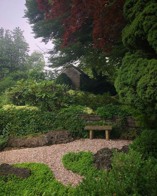 A serene garden scene featuring lush greenery, a stone structure, and a wooden bench surrounded by gravel paths.