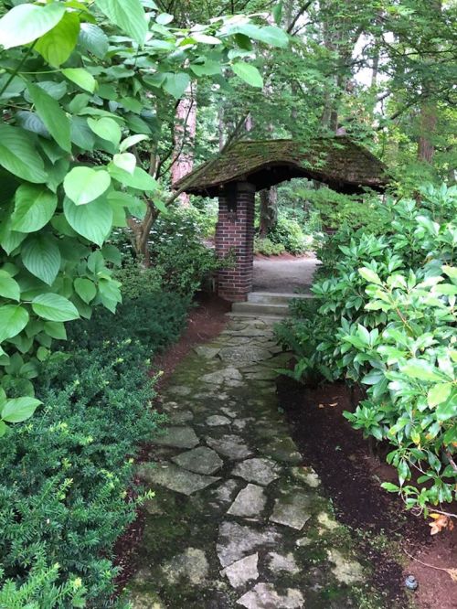 A stone pathway leads to a covered archway surrounded by lush greenery and trees.