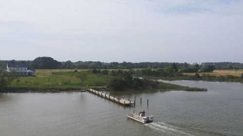 A boat glides across a calm river near a grassy shore and a wooden dock, with trees in the background under a clear sky.