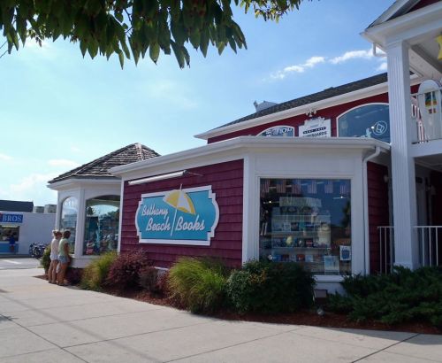 A charming bookstore with a red exterior, featuring a sign that reads "Bethany Beach Books" and surrounded by greenery.