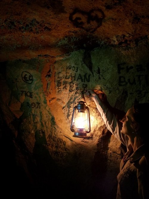A person holds a lantern in a dimly lit cave, illuminating rock walls with faint markings.