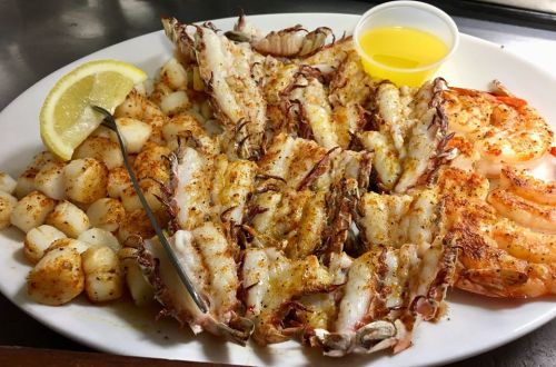 A plate of seasoned seafood featuring shrimp, lobster tail, and scallops, served with lemon and a small cup of butter.