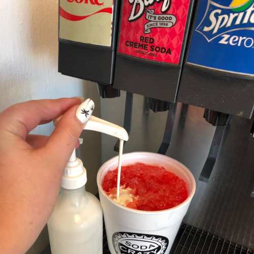 A hand pours cream into a cup of red soda at a beverage dispenser.