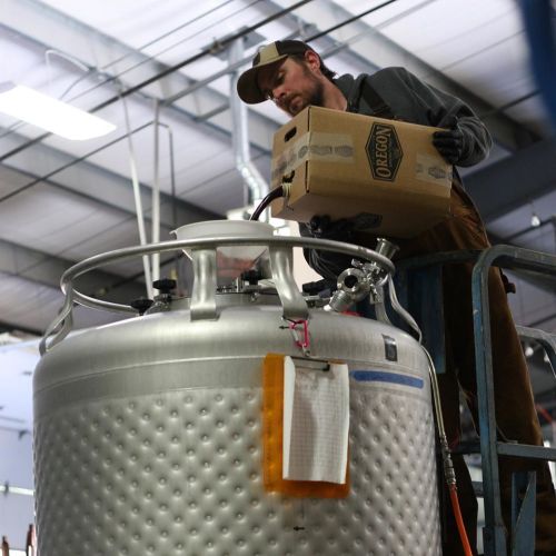 A man in a cap places a box on top of a large silver tank in a warehouse setting.