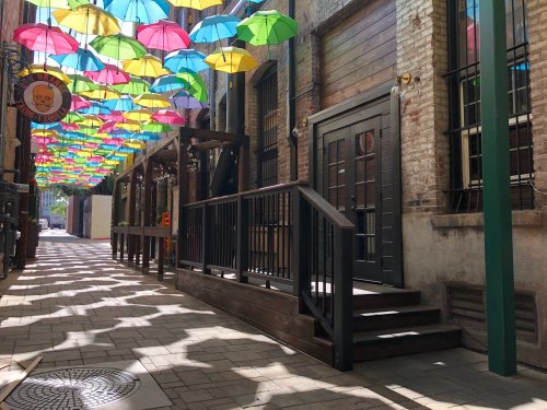Colorful umbrellas hang overhead in a charming alleyway, with a wooden deck and brick walls creating a vibrant atmosphere.