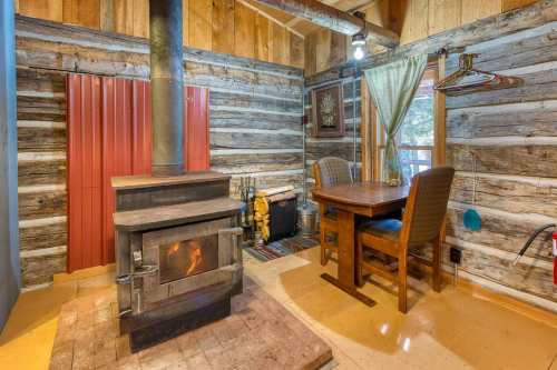 Cozy cabin interior featuring a wood stove, wooden walls, a small table, and a chair by a window.