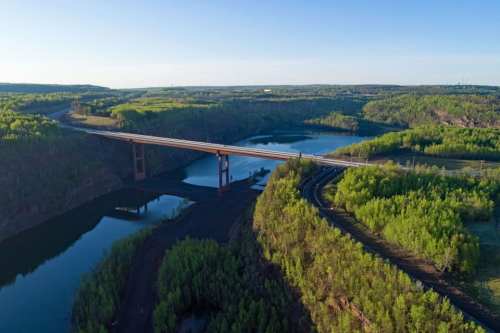 Aerial view of a bridge spanning a river, surrounded by lush green forests and rolling hills under a clear blue sky.