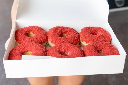 A person holding a white box containing six red velvet donuts with a crumb topping.