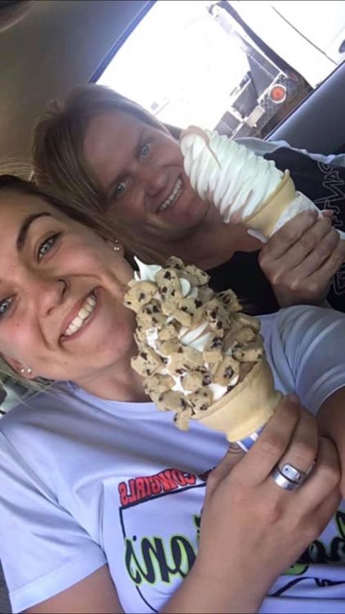 Two smiling women hold large ice cream cones, one with cookie pieces and the other with soft-serve, inside a car.