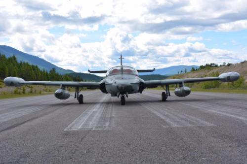 A military aircraft on a runway, surrounded by trees and mountains under a cloudy sky.