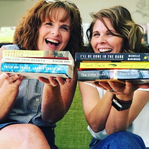 Two women hold up stacks of books, smiling and posing together in a cozy setting.
